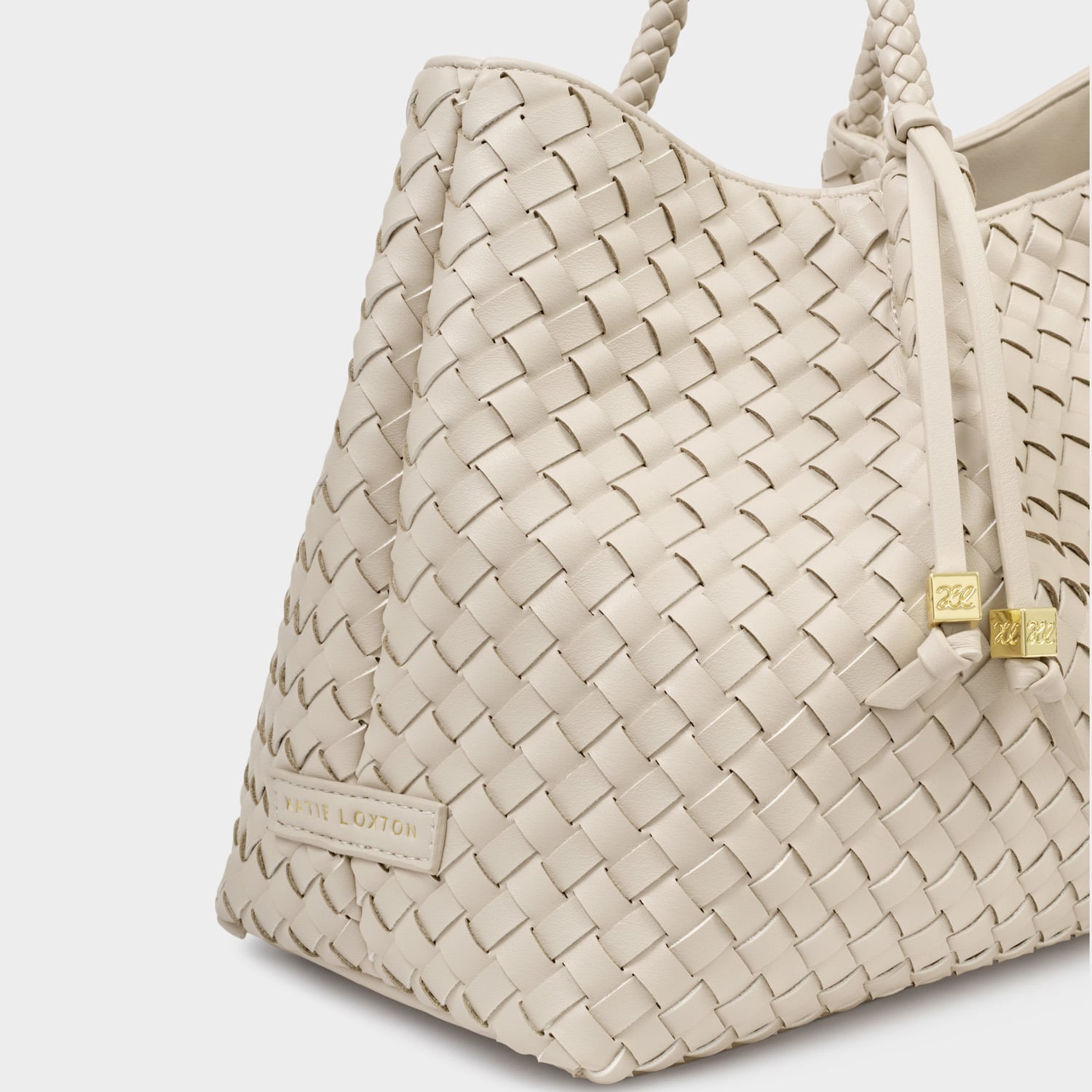 Photograph of a Katie Loxton cream-colored woven tote bag in a studio featuring a diagonal basketweave texture, braided handles, and iconic gold-tone initialed charms