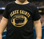 Photograph of a toddler at a football stadium wearing a black t-shirt with a distressed gold Geaux Saints graphic that features the New Orleans Superdome and skyline silhouette within a football shape