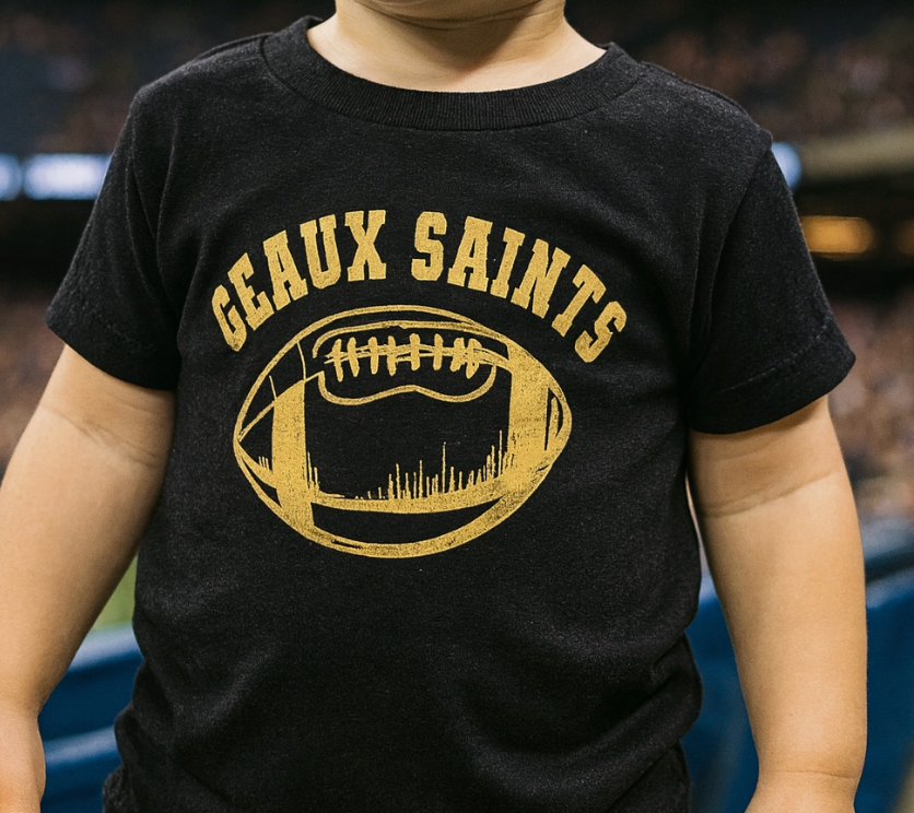 Photograph of a toddler at a football stadium wearing a black t-shirt with a distressed gold Geaux Saints graphic that features the New Orleans Superdome and skyline silhouette within a football shape