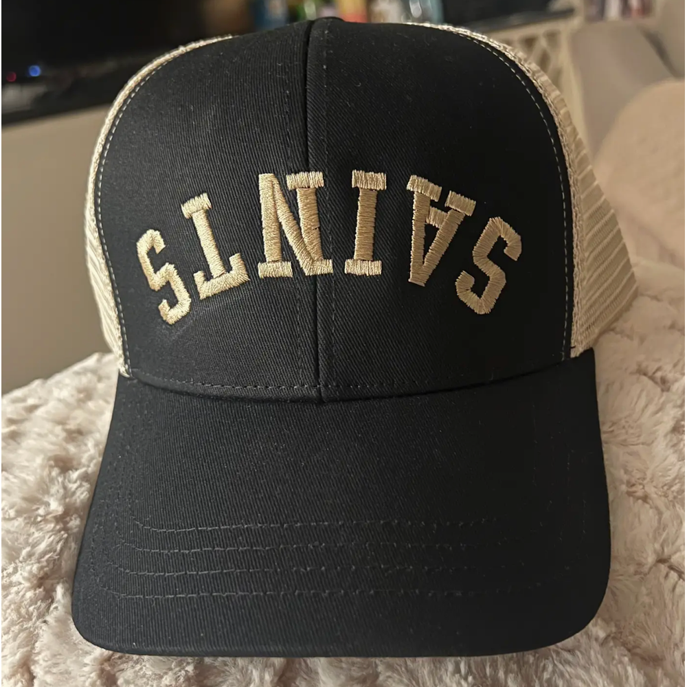 Photograph of a black and tan trucker hat featuring arched tan SAINTS embroidery on the front panel resting on a textured cream plush surface with a breathable mesh back and a curved brim with parallel stitching