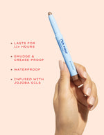 A female hand holds a light blue Alleyoop 11th Hour Eyeshadow Stick with a bronze tip, highlighting its 11+ hour wear, smudge and crease-proof, waterproof, and jojoba oil infused benefits against a soft peach background