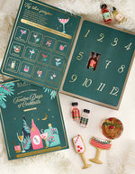 A Twelve Days of Cocktail Advent calendar set arranged on a white faux-fur surface. The set includes a dark teal numbered box with a window showing one bottle, a front cover illustration, a recipe card, and several small cocktail-themed bottles and decorative accessories.