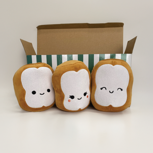Three plush beignets with faces in front of a cardboard box on a light background