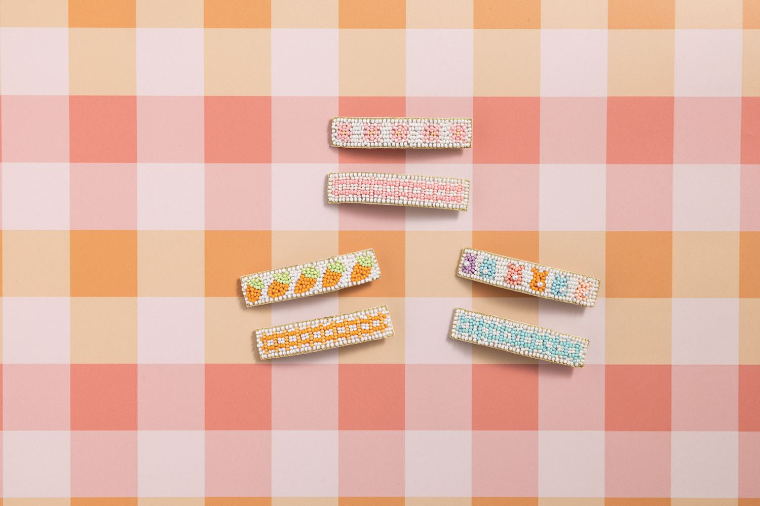 Six colorful beaded hair clips featuring pastel carrot, flower, BUNNY, and geometric checkerboard patterns arranged on a light orange and pink gingham background