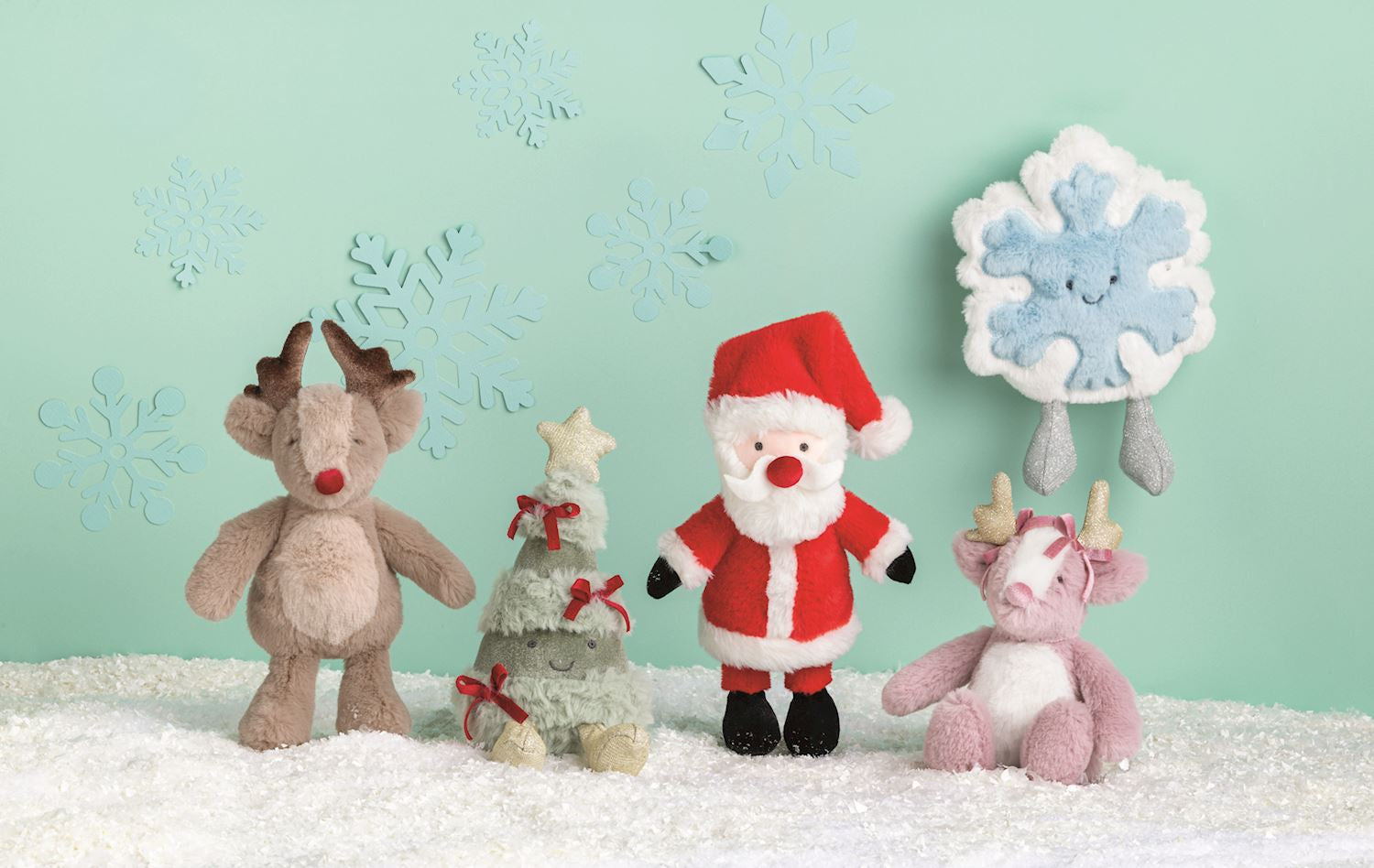 Plush reindeer, Christmas tree, Santa Claus, and snowflake toys on a snowy surface against a light green background