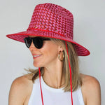 Person wearing a pink woven sun hat with a red strap on a plain background