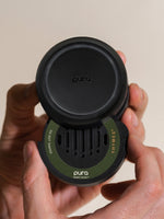 Photograph of a Pura Smart Scent fragrance vial held by hands against a neutral background featuring a matte black circular body, a moss green Thymes brand label, and vertical air vents