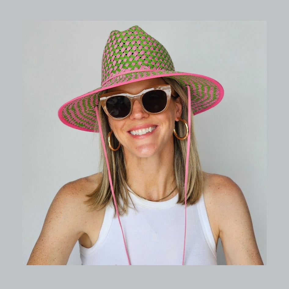 A person wearing a green and pink woven lifeguard hat with a white tank top and sunglasses