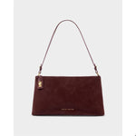Product photograph by Katie Loxton of a deep burgundy faux suede shoulder bag in a studio setting featuring a gold-tone turn-lock tag, thin adjustable strap, and minimalist brand lettering