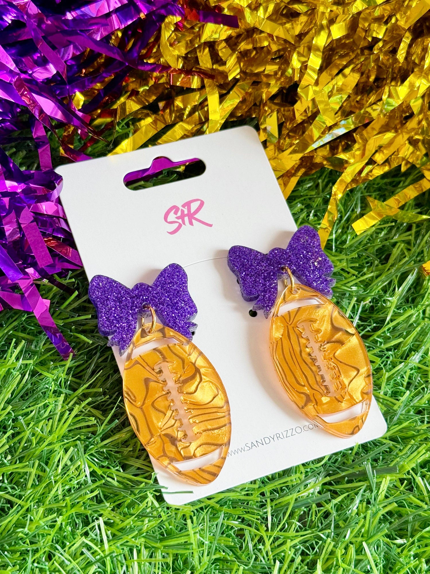 Acrylic earrings by SMR (Sandy Rizzo) featuring glittery purple bow studs and gold football drops with embossed laces on a white display card set against artificial green grass with shimmering purple and gold tinsel