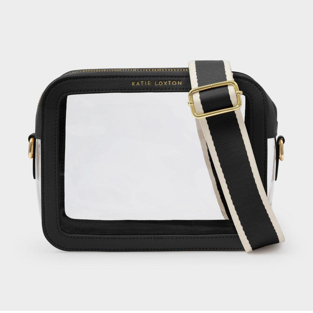 Product photograph by Katie Loxton of a clear crossbody stadium bag on a neutral background featuring black vegan leather trim, gold-tone logo hardware, and a wide striped canvas strap