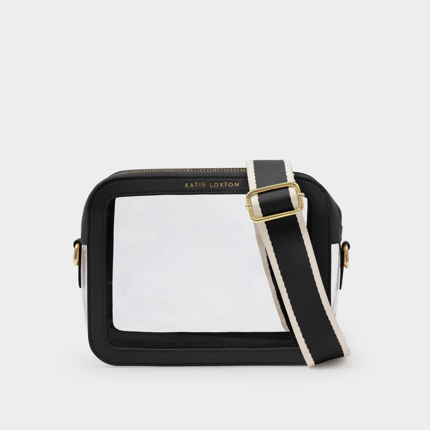 Product photograph by Katie Loxton of a clear crossbody stadium bag on a neutral background featuring black vegan leather trim, gold-tone logo hardware, and a wide striped canvas strap