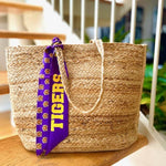 Purple twilly scarf with "TIGERS" in gold letters and tiger faces tied to the handle of a woven jute tote
