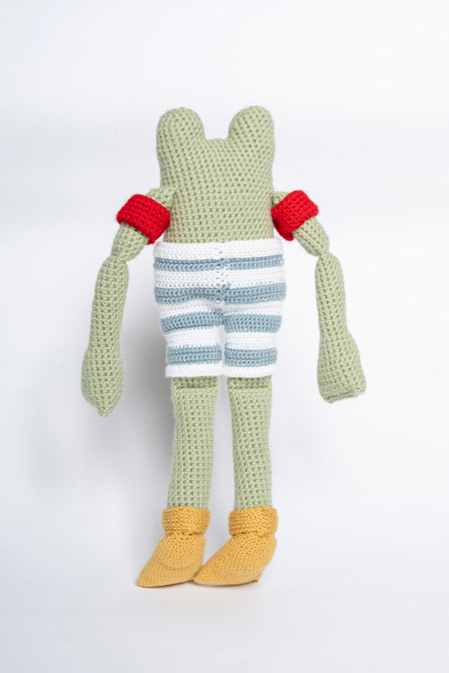 Crochet light green frog character, viewed from the back, wearing red armbands, blue and white striped shorts, and yellow booties on a plain white background