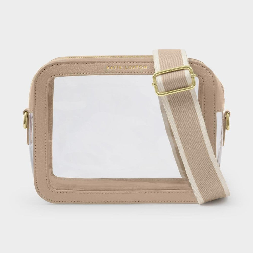 Product photography of a Katie Loxton beige and clear crossbody bag featuring a transparent PVC panel, gold logo hardware, and a wide striped adjustable shoulder strap on a neutral studio background