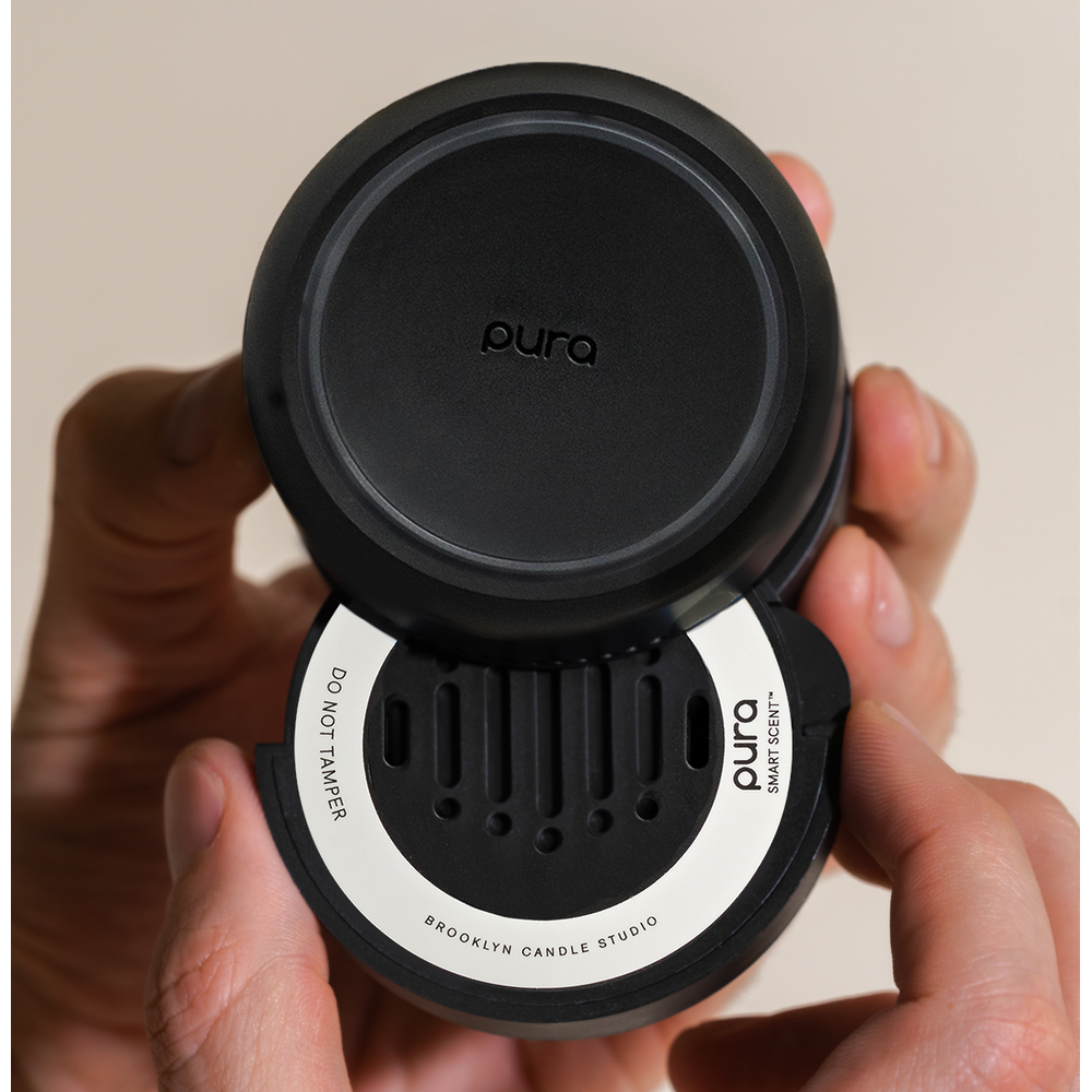 Commercial photograph for Pura of a matte black Smart Scent fragrance refill held in hands featuring a white Brooklyn Candle Studio label and vertical slotted vents