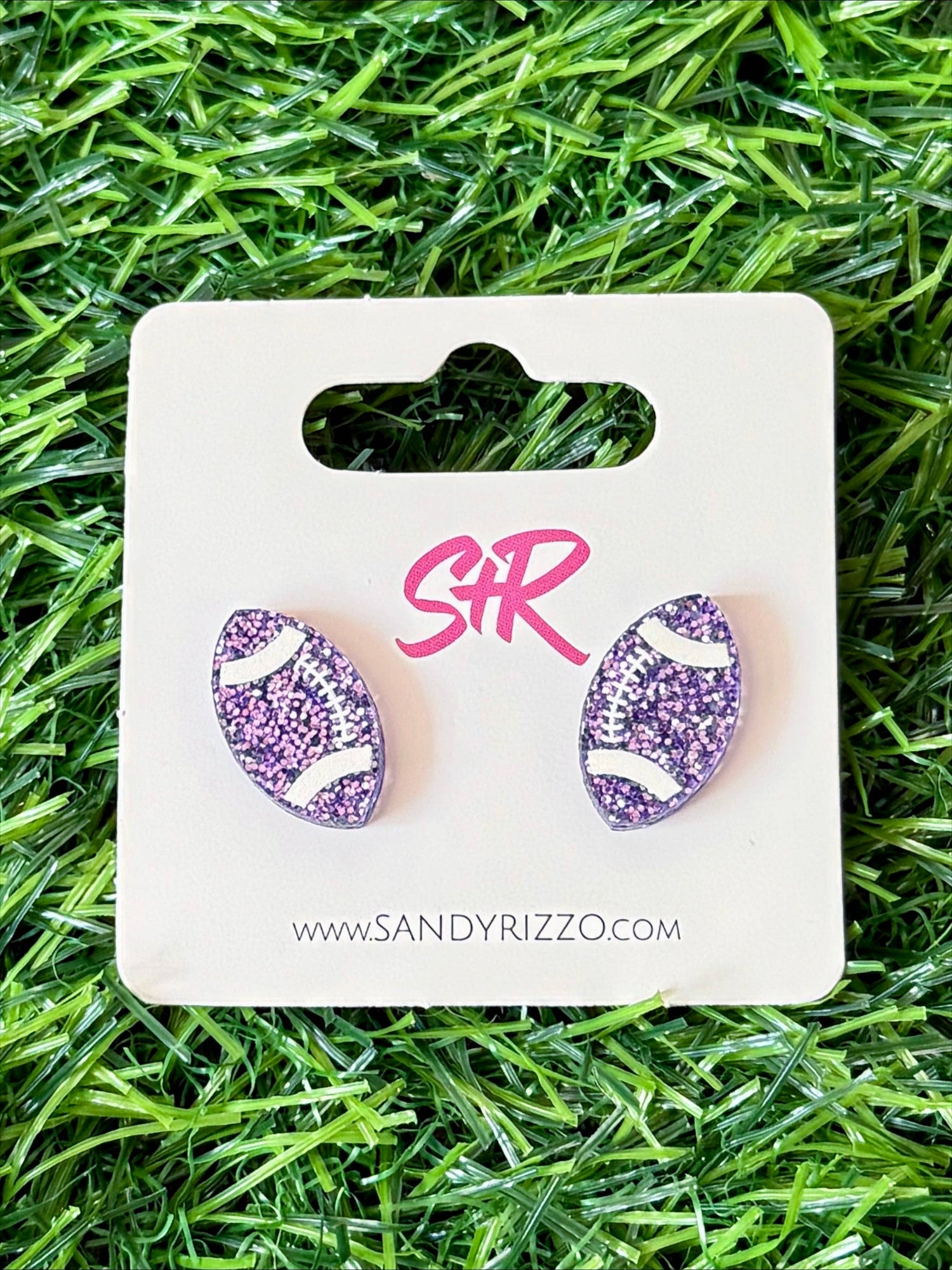 Sandy Rizzo fashion jewelry, a pair of glittery purple football stud earrings with white lacing on a white display card, set against green artificial turf with a pink SR logo and brand website