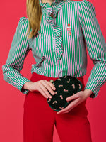 Studio fashion photograph of a model wearing a pink and green striped ruffled shirt with a nutcracker embroidery and red trousers holding a dark green velvet clutch with candy cane motifs against a bright pink background