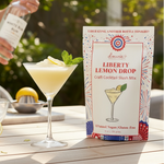Liberty Lemon Drop cocktail mix box with a cocktail and lemon on a table outdoors.