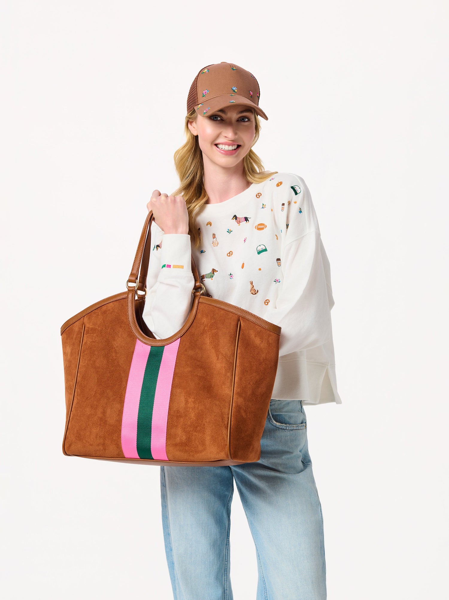 Woman wearing a brown baseball cap and white embroidered sweatshirt holding a brown suede tote bag with striped detail