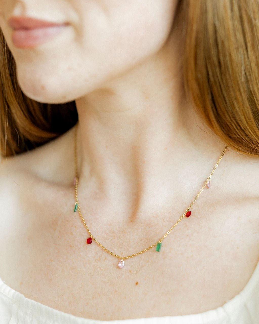 A woman's décolletage features a delicate gold chain necklace adorned with vibrant red, pink, and green gemstone charms