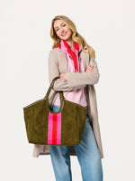 Woman wearing a beige coat and pink sweater holding an olive green suede tote bag with pink and red stripes