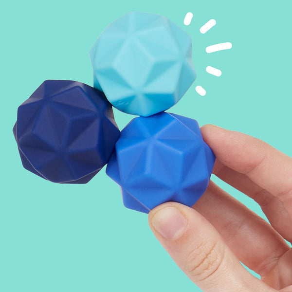 Digital photograph of three faceted geometric fidget toys in shades of navy, royal, and sky blue held by a hand against a solid turquoise background with white illustrative sparkle accents