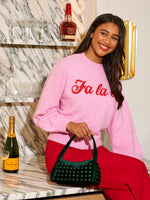Digital photograph of a smiling woman wearing a pink Fa la knit sweater and red trousers sitting on a marble bar counter featuring bottles of Veuve Clicquot and Maker's Mark while holding a dark green studded velvet handbag