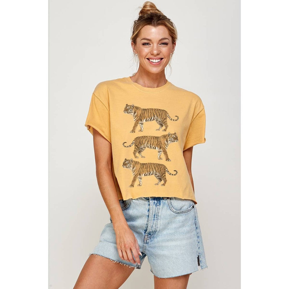 Digital photograph of a smiling woman in a minimalist studio wearing a mustard yellow crop top with three stacked tigers and light wash frayed denim shorts