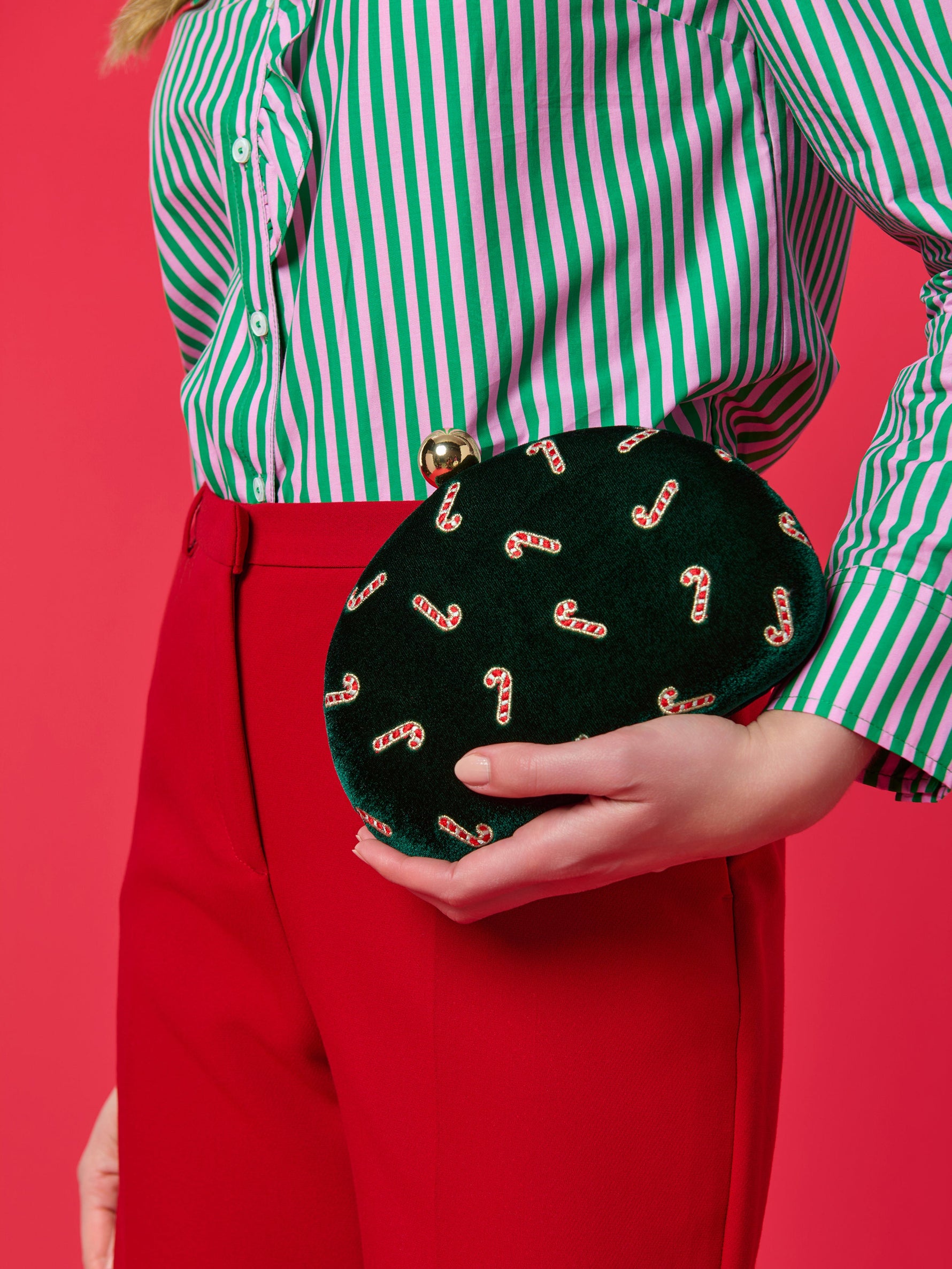 Digital photograph of a festive green velvet clutch with candy cane embroidery and a gold ball clasp held by a person wearing a pink and green striped shirt and red trousers against a vibrant pink background