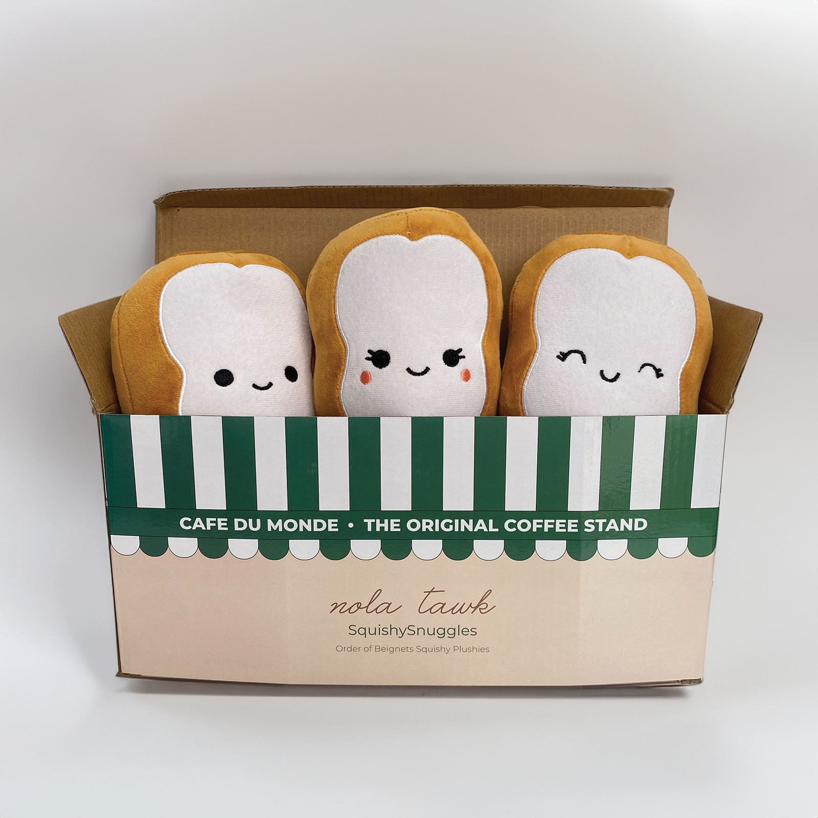 Three plush toys resembling beignets with faces in a box labeled 'Cafe du Monde'.