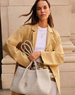 Fashion photograph of a woman standing against a classical stone building facade wearing a tan oversized blazer and holding a large white woven leather tote bag with thick braided rope handles and gold jewelry accents