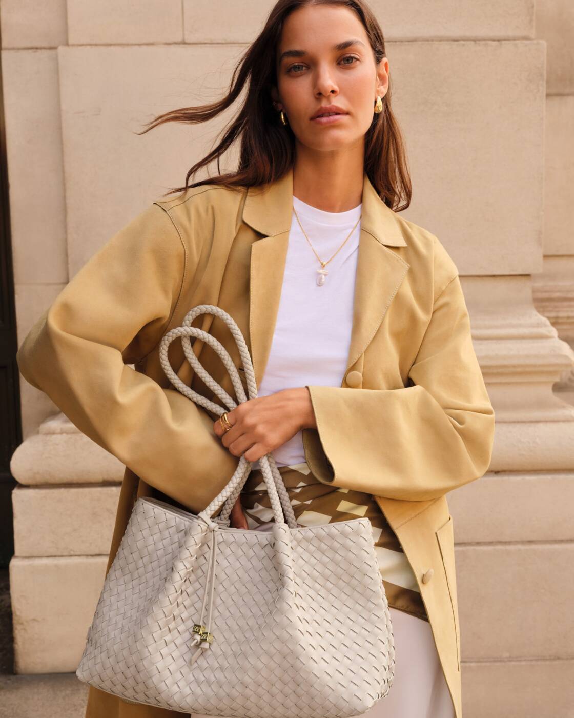 Fashion photograph of a woman standing against a classical stone building facade wearing a tan oversized blazer and holding a large white woven leather tote bag with thick braided rope handles and gold jewelry accents