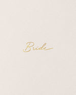 Close-up photograph of a minimalist wedding journal featuring the word Bride in shimmering gold foil cursive script centered on a fine-grained, off-white linen texture