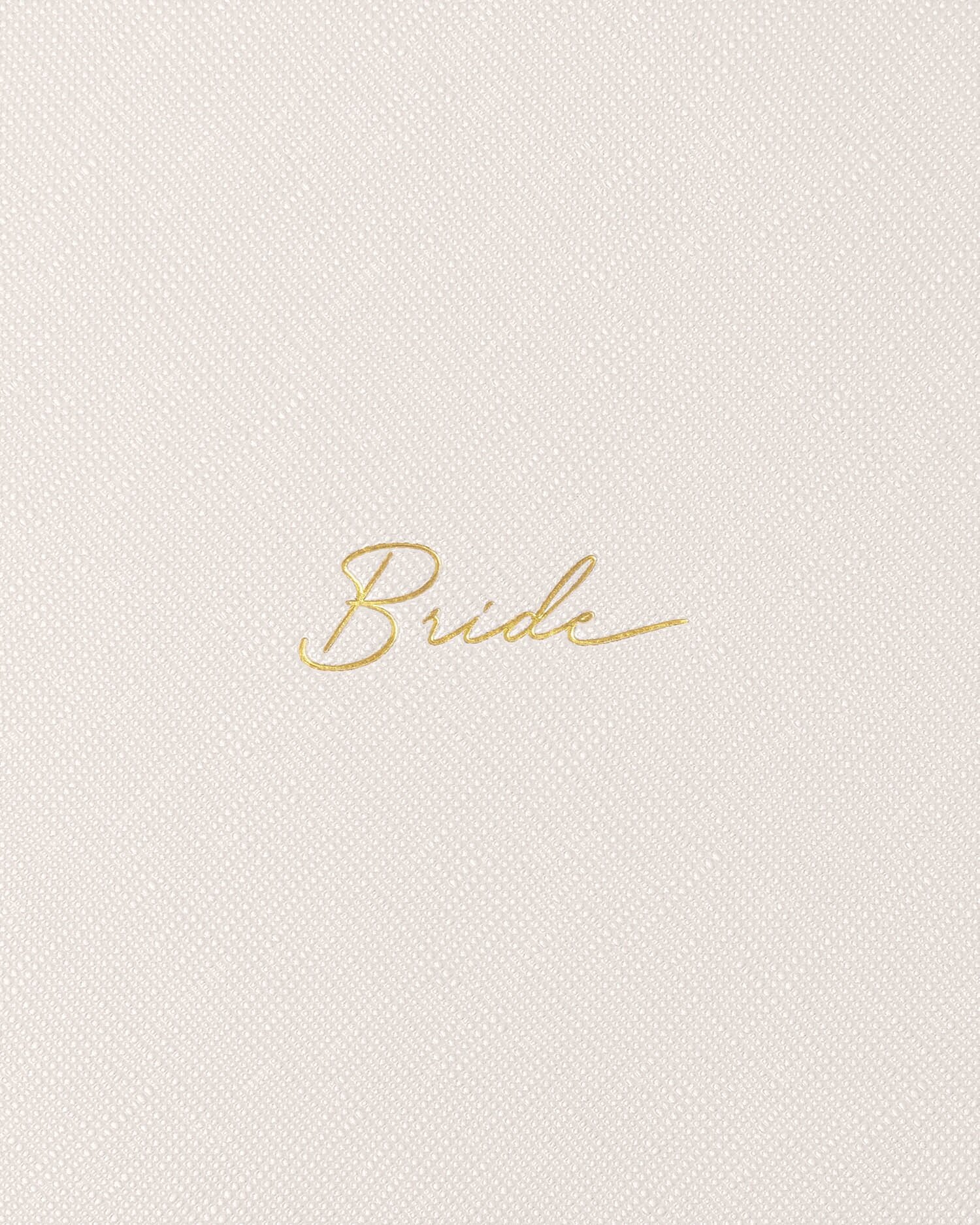 Close-up photograph of a minimalist wedding journal featuring the word Bride in shimmering gold foil cursive script centered on a fine-grained, off-white linen texture
