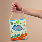 Digital photograph of a Floss & Rock triceratops shaped puzzle box held by a hand against a tan background featuring a vibrant orange and white striped rope handle, a blue and white striped top border, and colorful dinosaur fossil and footprint illustrations