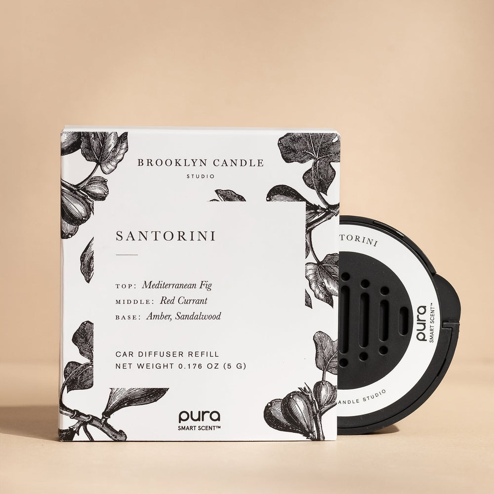 Product photograph by Brooklyn Candle Studio and Pura of a Santorini car diffuser refill kit featuring a white box with black botanical fig illustrations and a circular black smart scent pod against a neutral beige studio background
