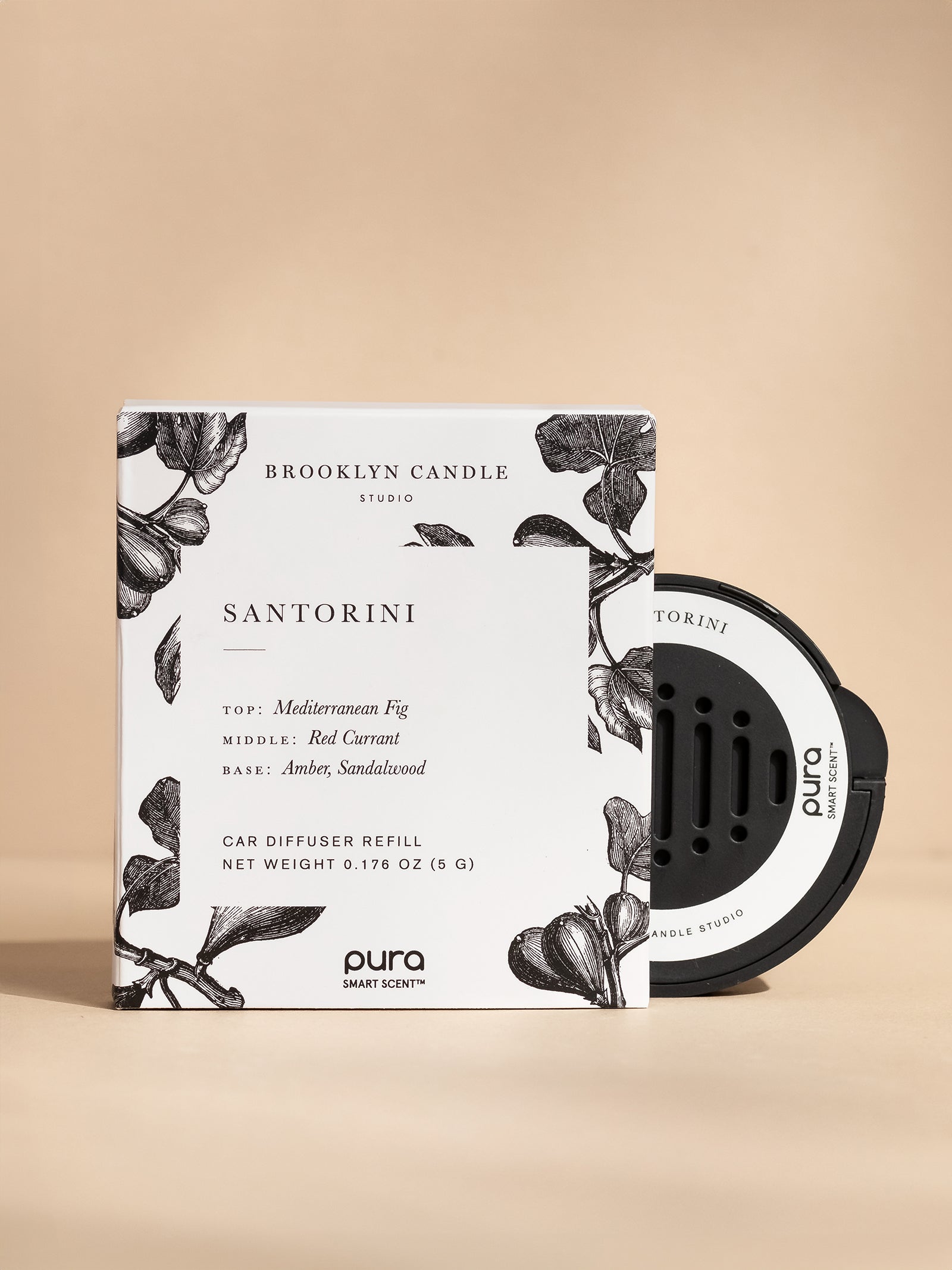 Product photograph by Brooklyn Candle Studio and Pura of a Santorini car diffuser refill kit featuring a white box with black botanical fig illustrations and a circular black smart scent pod against a neutral beige studio background