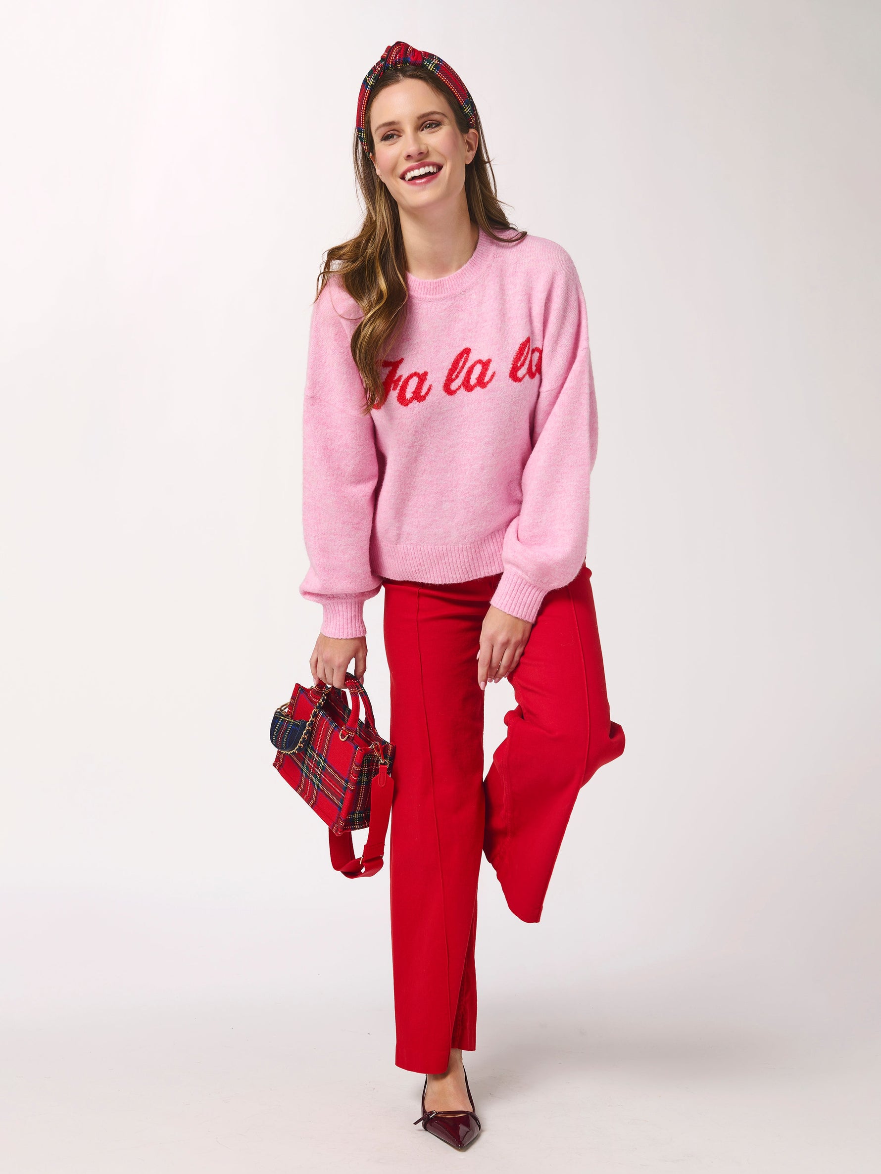 Studio photograph of a woman modeling a festive holiday outfit in a white studio featuring a pink sweater with red fa la la script, red wide-leg trousers, and a red and navy plaid headband with a matching tartan handbag