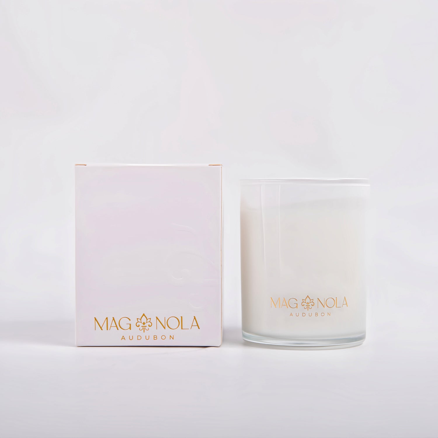 Studio photograph by Magnola Audubon of a luxury scented candle and packaging box set against a minimalist white background featuring gold foil lettering, a stylized fleur-de-lis emblem, and a frosted glass vessel filled with creamy white wax