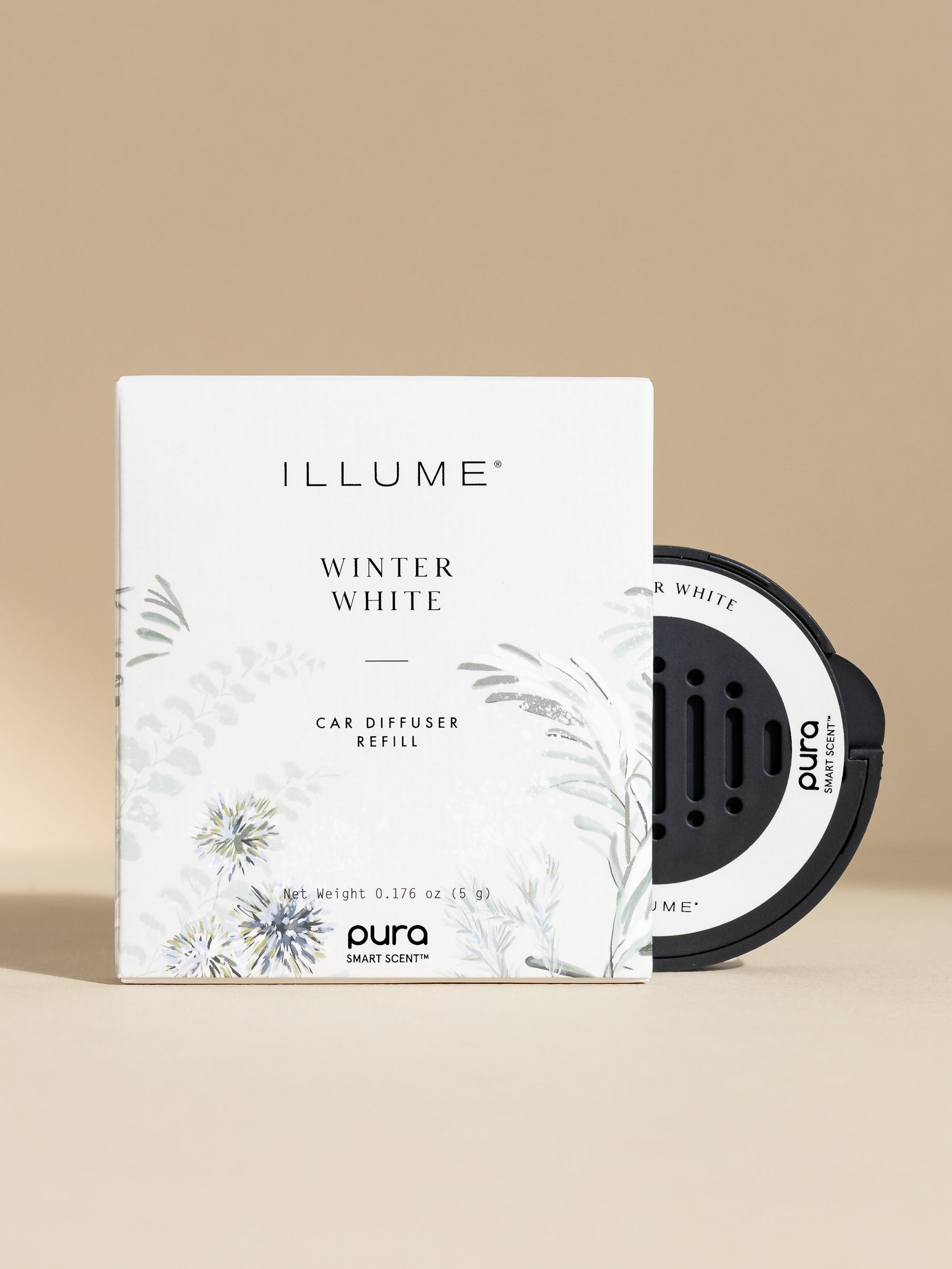 Commercial product photography by Illume and Pura of a Winter White car diffuser refill featuring a white box with delicate sage green botanical illustrations and a circular black scent cartridge with perforated vents against a neutral beige backdrop