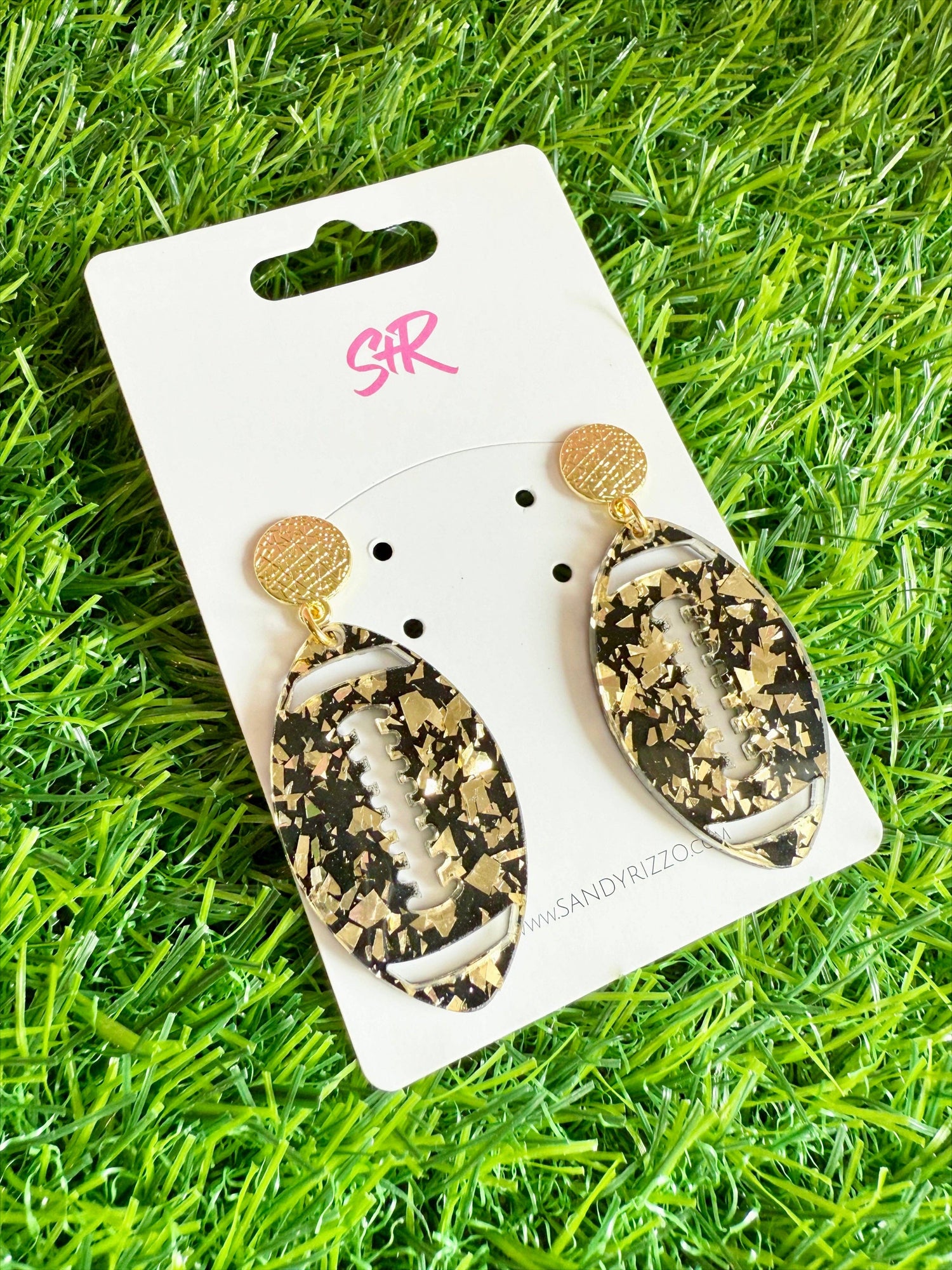 Sandy Rizzo resin and gold-tone football-shaped earrings featuring black resin with gold foil flakes and textured gold stud tops displayed on a white S+R branded card against a vibrant green artificial turf background