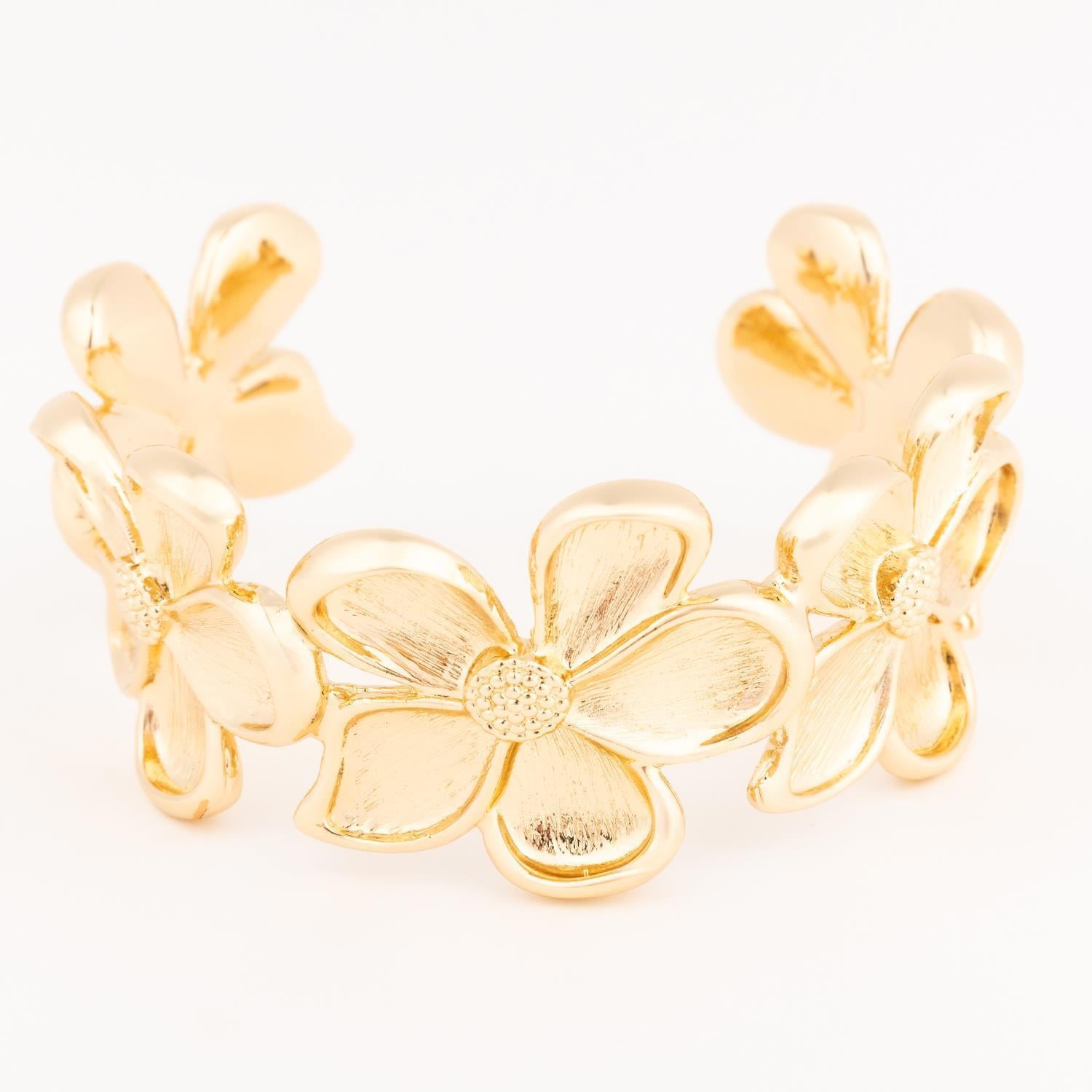 Polished gold-tone metal floral cuff bracelet on a white background featuring sculpted five-petal flowers with textured stippled centers and a satin-brushed finish on the petals