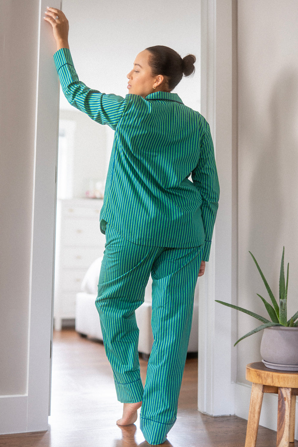 Photograph of a woman wearing striking green and blue striped pajamas with her hair in a bun standing barefoot in a bright doorway looking over her shoulder with an aloe vera plant on a light wooden stool nearby
