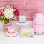 Pink tea set with a teapot, cup, and tea canister against a pink brick wall.