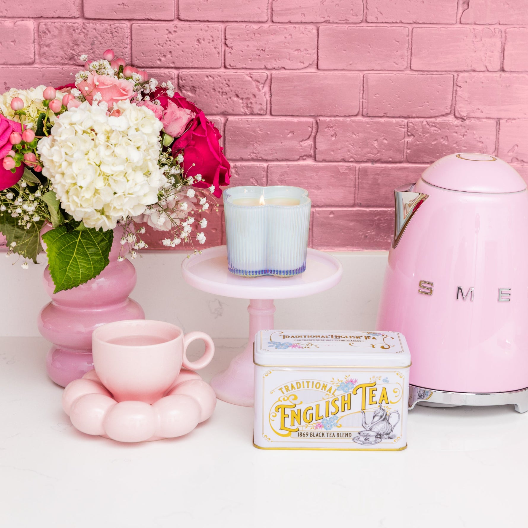 Pink tea set with a teapot, cup, and tea canister against a pink brick wall.