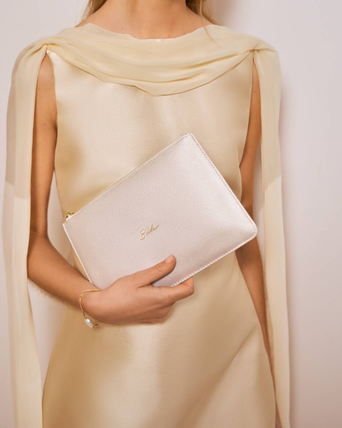 Digital photograph of a shimmering pearlescent Katie Loxton bridal clutch held by a woman in a champagne satin dress featuring gold cursive Bride script and delicate sheer draped sleeves