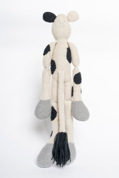 Crochet stuffed cow featuring an off-white body with black spots, long limbs with grey feet, and a black tassel tail against a plain white background