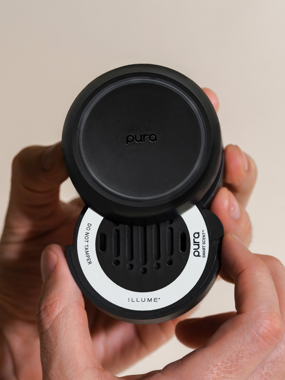 Photograph of a Pura Smart Scent device held in a person's hands, featuring the Illume fragrance refill with its white circular label, vertical ventilation slits, and the embossed Pura logo against a neutral beige background