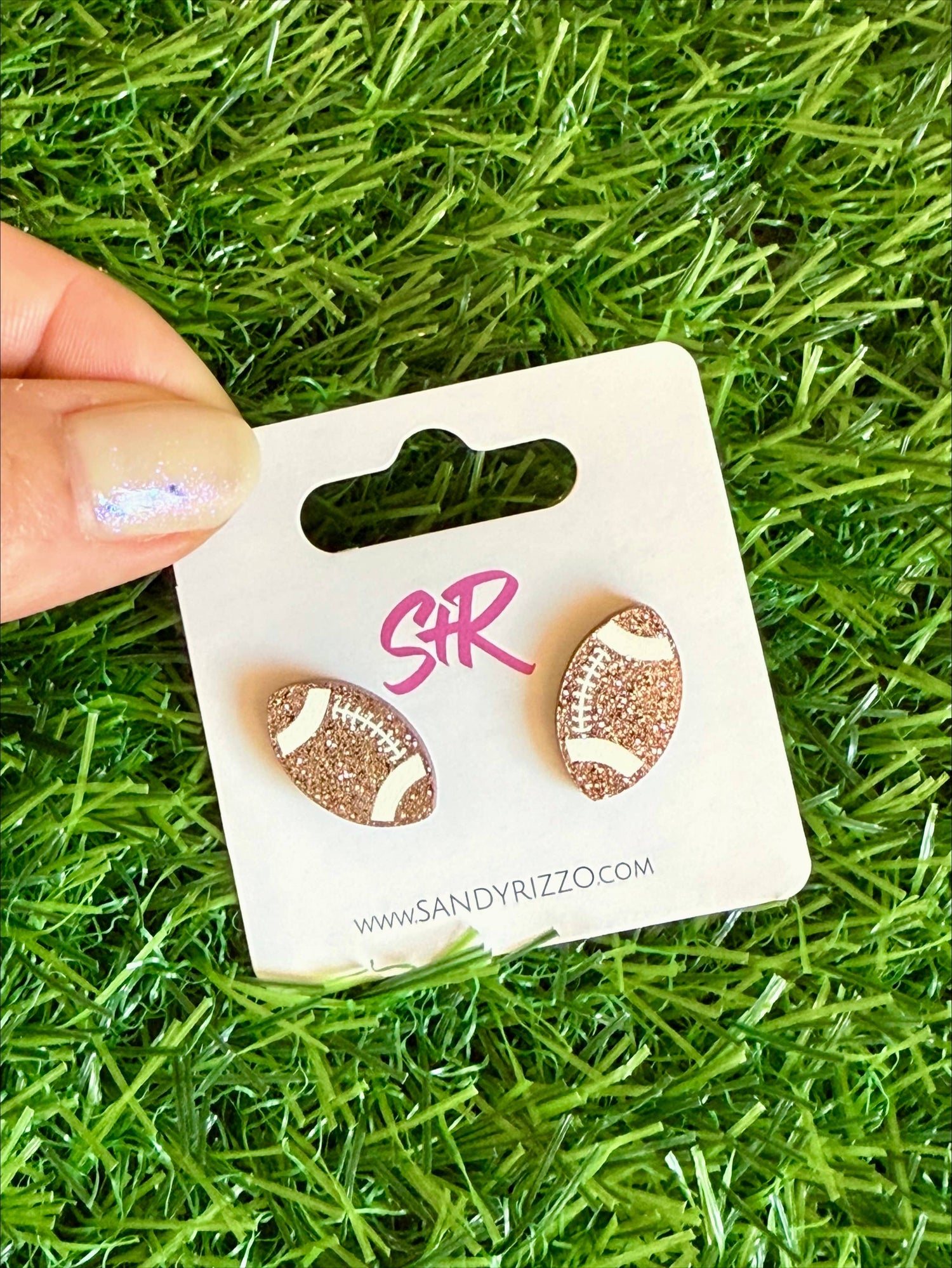 Sandy Rizzo earrings featuring two glittery rose gold football stud earrings with white lacing on a white display card with a pink SR logo, held by a hand with iridescent nail polish over green artificial turf